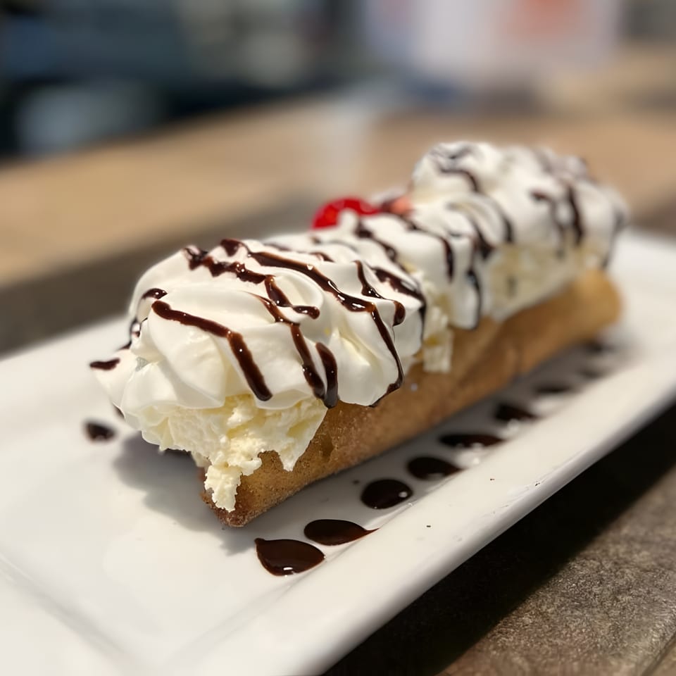 A long churro topped with whipped cream, chocolate drizzle, and a cherry on a white plate.