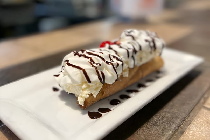 A long churro topped with whipped cream, chocolate drizzle, and a cherry on a white plate.