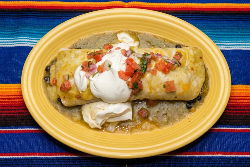 A large burrito covered in green chili, cheese, sour cream, and pico de gallo on a yellow plate.
