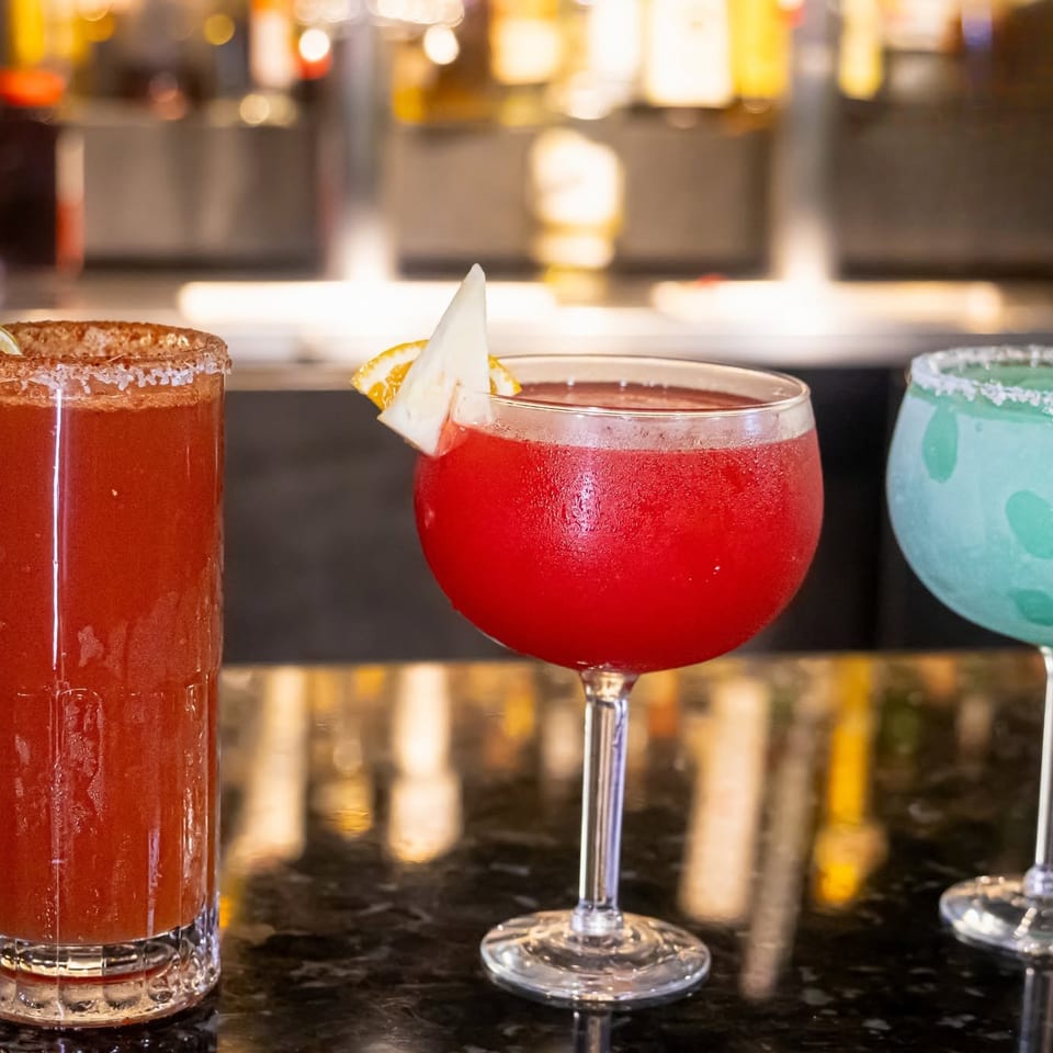 Three colorful cocktails, including a red michelada, a red margarita, and a blue margarita, on a bar.