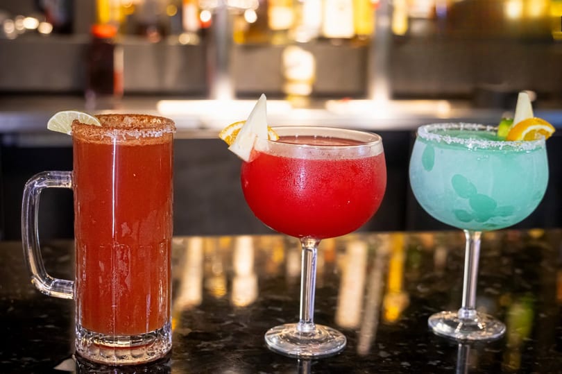 Three colorful cocktails, including a red michelada, a red margarita, and a blue margarita, on a bar.