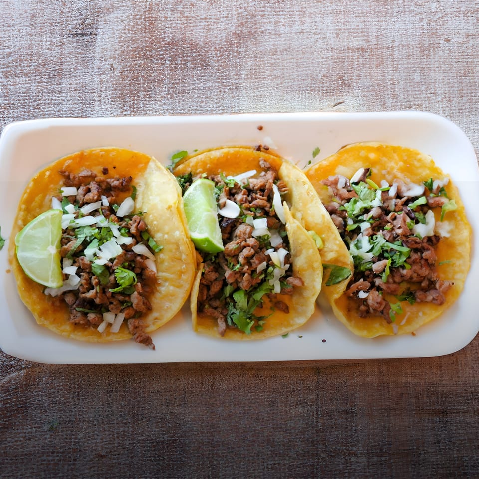 Three street tacos with meat, onions, cilantro, and lime wedges on a white plate.
