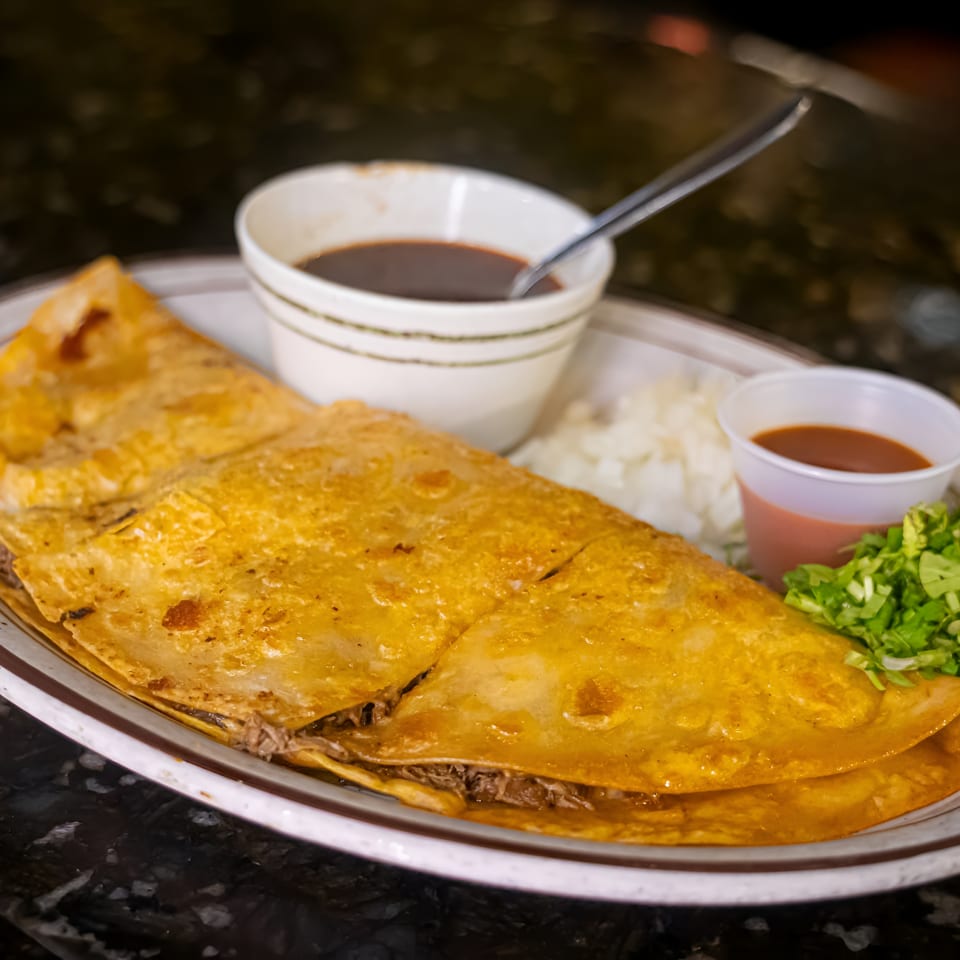 A large, golden-brown quesadilla filled with meat, served with dipping sauces, onions, and lettuce.