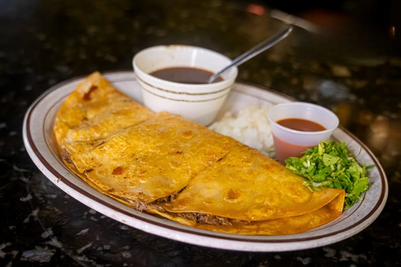 A large, golden-brown quesadilla filled with meat, served with dipping sauces, onions, and lettuce.